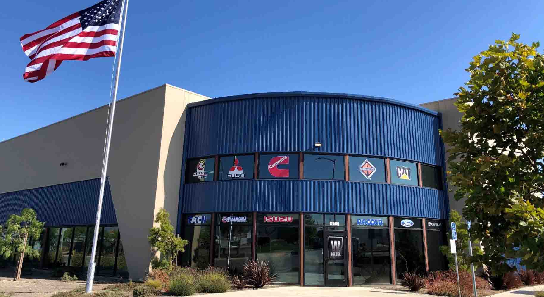 Watsonville Diesel headquarters building - front view. An American flag blows in the wind. The building is blue and tan. It's new construction.